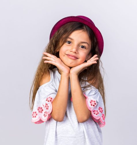pleased little caucasian girl with purple party hat putting hands on chin isolated on white background with copy space
